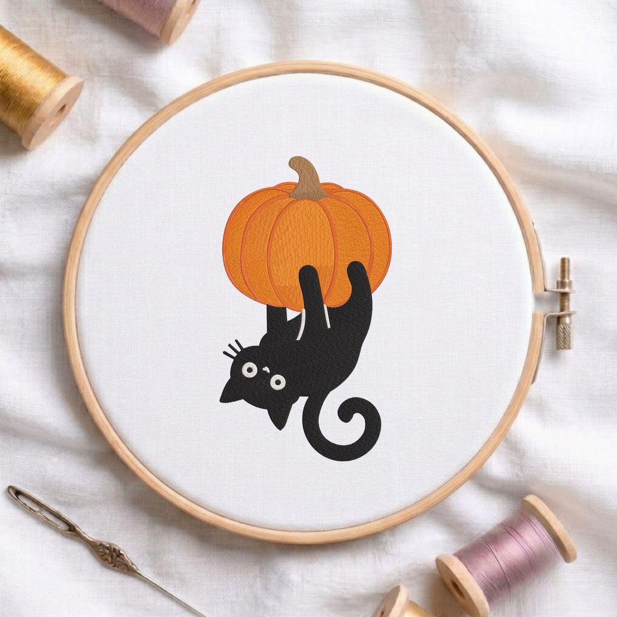 Mischievous black cat hanging upside down from a pumpkin Halloween machine embroidery design shown in a wooden embroidery hoop on white fabric with thread spools and a needle