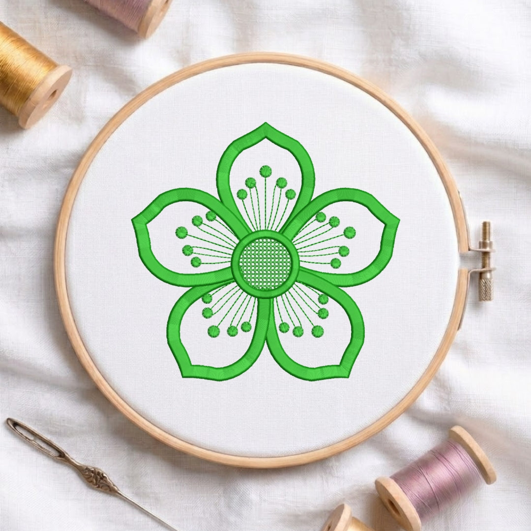 A circular embroidery hoop holding white fabric with a bright green flower design stitched in the center. The hoop is photographed on a white fabric surface with spools of thread and a needle around it.