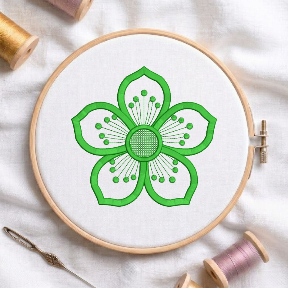 A circular embroidery hoop holding white fabric with a bright green flower design stitched in the center. The hoop is photographed on a white fabric surface with spools of thread and a needle around it.