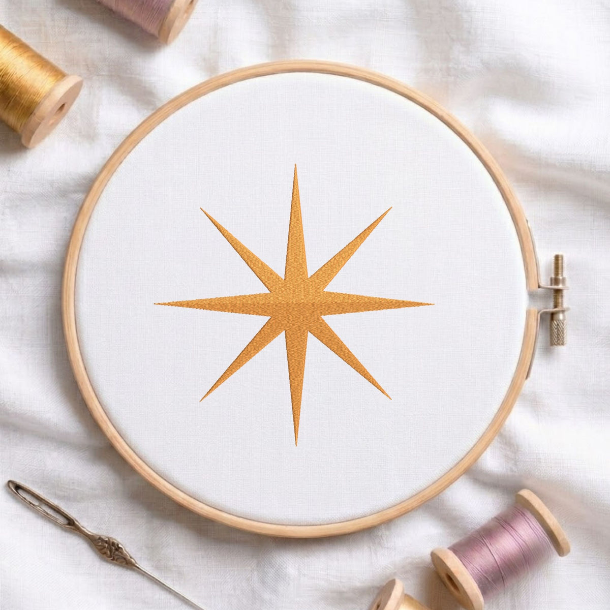 Golden starburst machine embroidery design displayed in a wooden hoop on white fabric with thread spools and a needle
