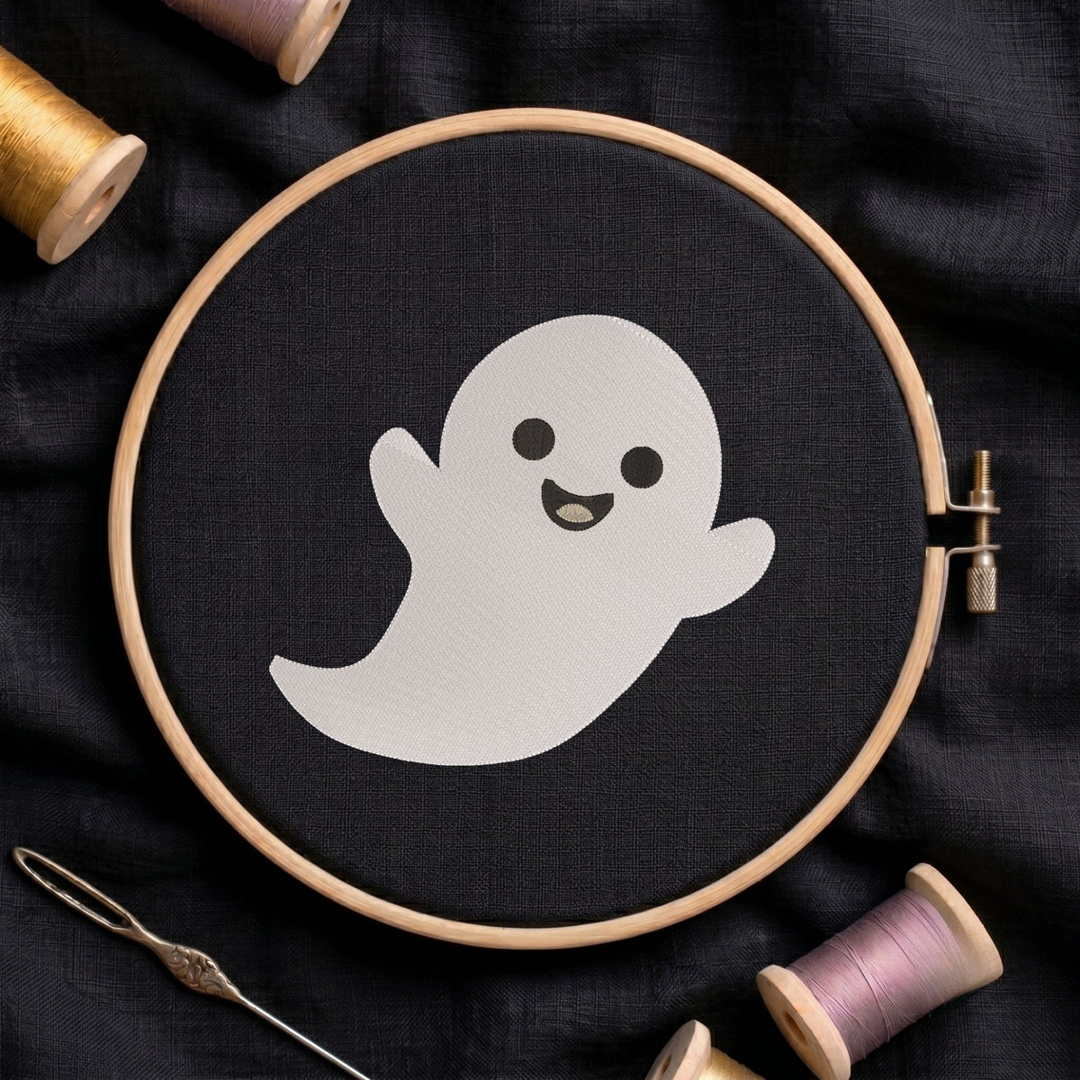 Cute smiling ghost machine embroidery design displayed inside a round wooden hoop on black fabric alongside thread spools and a needle.