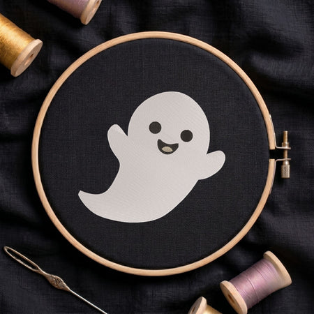 Cute smiling ghost machine embroidery design displayed inside a round wooden hoop on black fabric alongside thread spools and a needle.