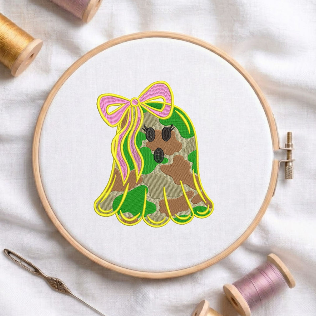  Cute camo ghost with a pink bow machine embroidery design displayed in a wooden hoop on white fabric with thread spools and a needle   