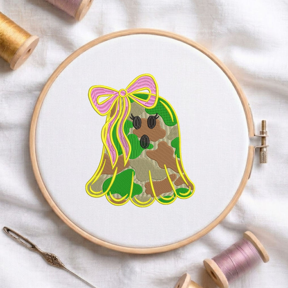 Cute camo ghost with a pink bow machine embroidery design displayed in a wooden hoop on white fabric with thread spools and a needle   