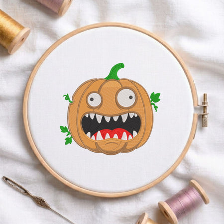 Funny spooky pumpkin with sharp teeth and wide eyes machine embroidery design displayed in a wooden hoop on white fabric with thread spools and a needle