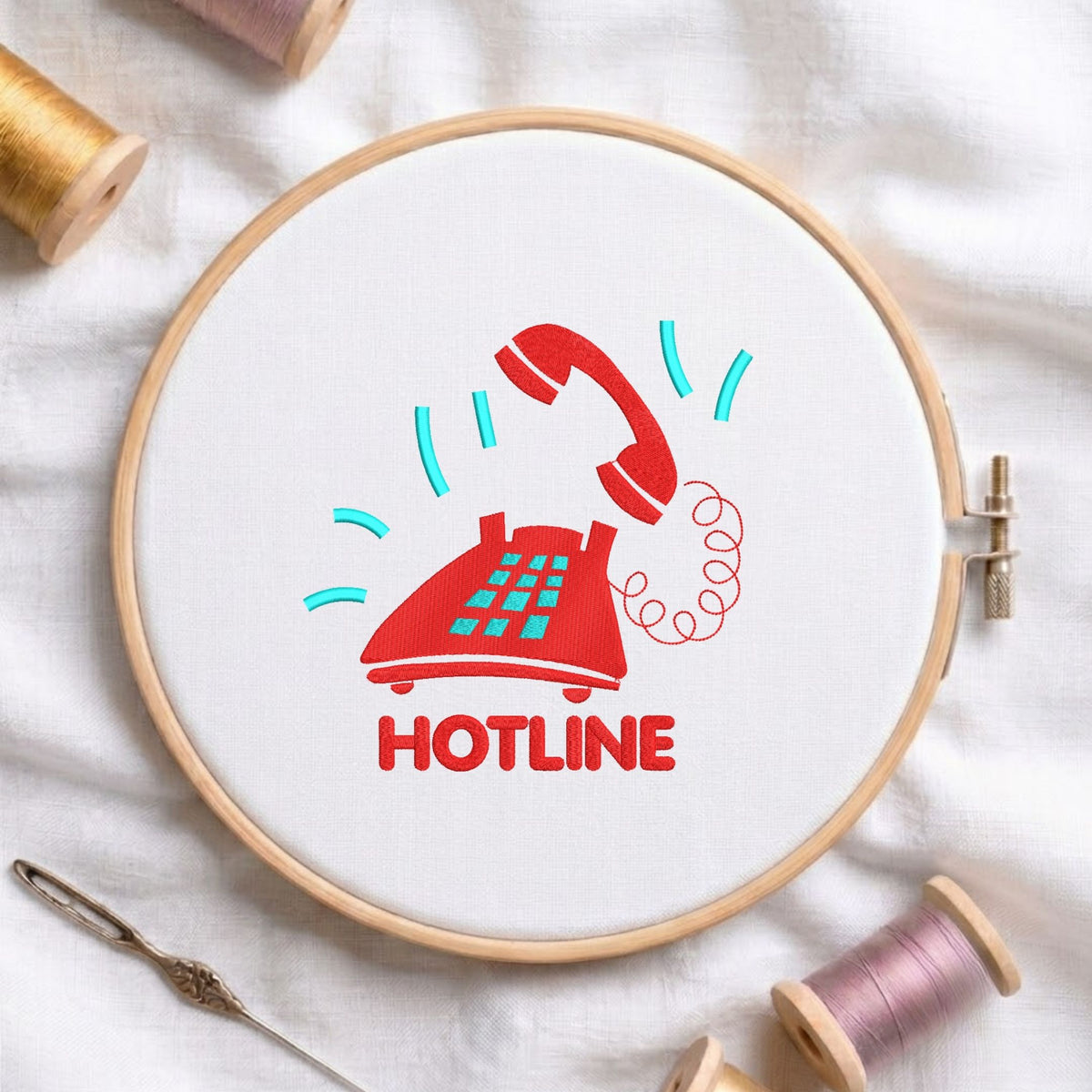 Red vintage telephone and "HOTLINE" text machine embroidery design displayed in a wooden hoop on white fabric with thread spools and a needle   