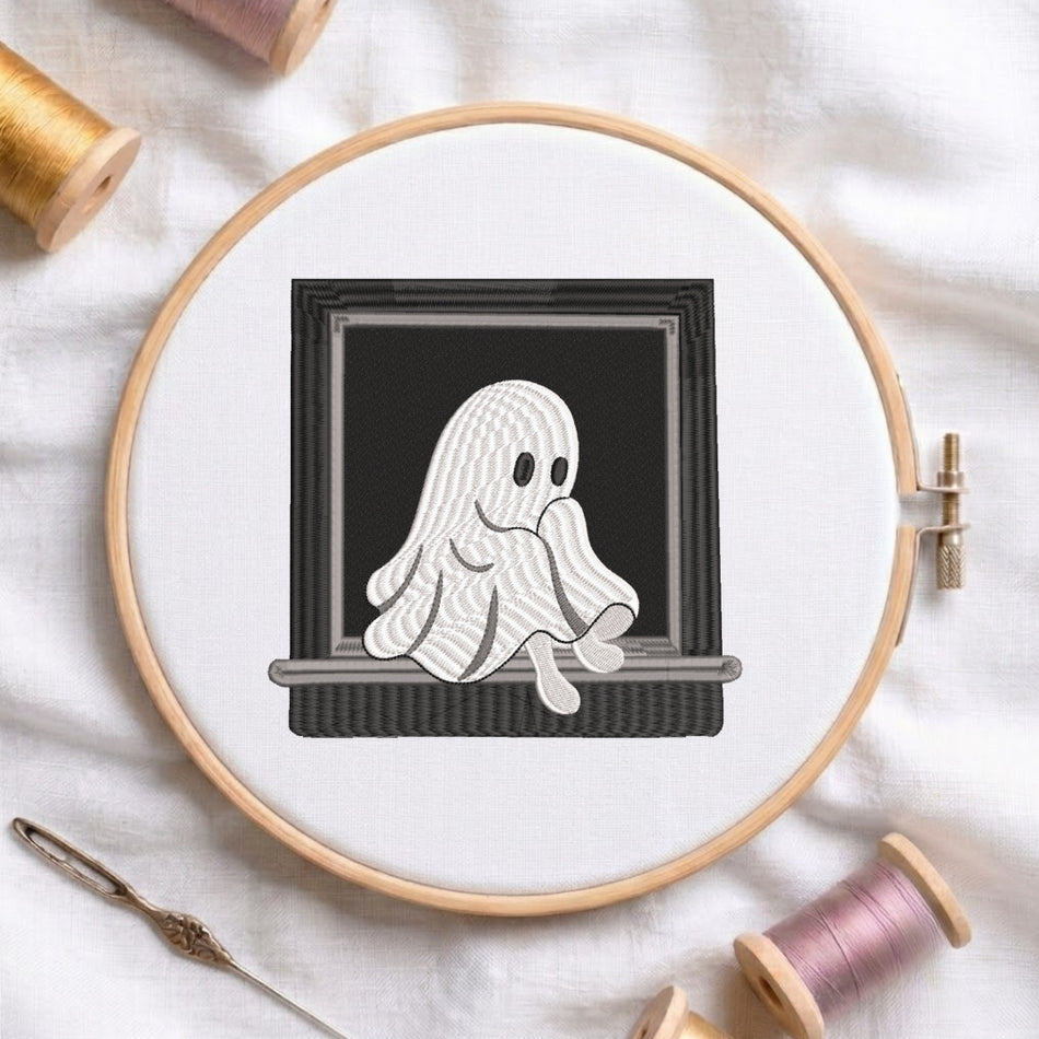 A circular embroidery hoop on white fabric, featuring a black-and-white stitched design of a ghost sitting in a chair. Spools of thread and a needle are arranged around the hoop.
