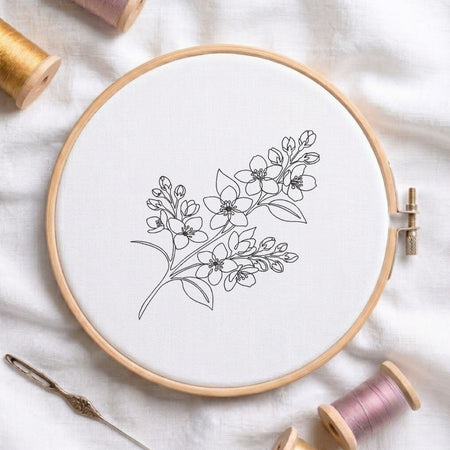 A round wooden embroidery hoop holding white fabric with a delicate black floral line drawing, styled on a white cloth with thread spools and a needle around it.