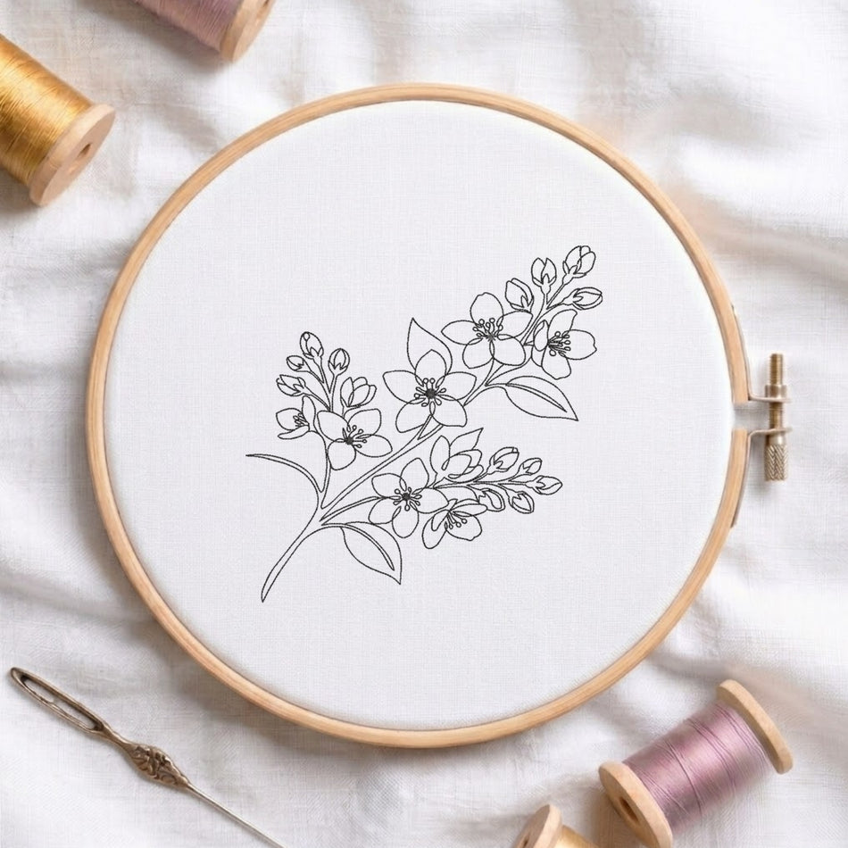 A round wooden embroidery hoop holding white fabric with a delicate black floral line drawing, styled on a white cloth with thread spools and a needle around it.