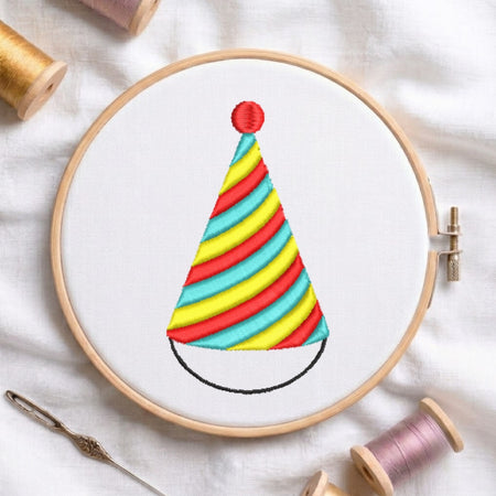 The party hat embroidery design secured in a wooden embroidery hoop, displayed with embroidery thread spools and a needle on a white fabric background.