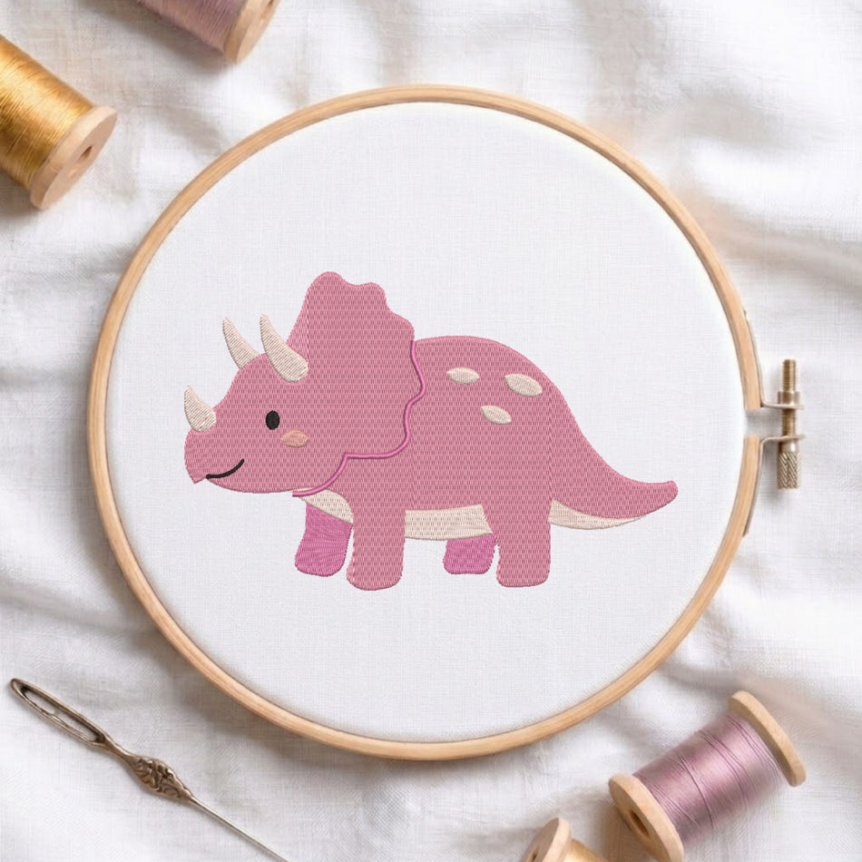 A pink embroidered Triceratops dinosaur design inside a wooden embroidery hoop, surrounded by spools of thread and a needle on a white fabric background.