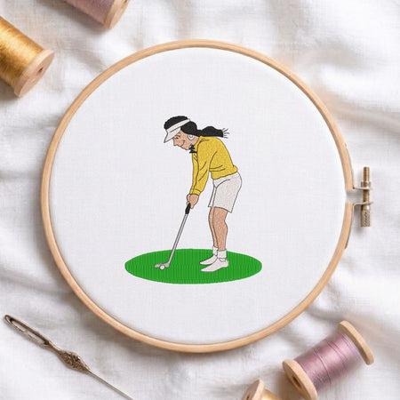 Woman playing golf machine embroidery design displayed in a wooden hoop on white fabric with thread spools and a needle