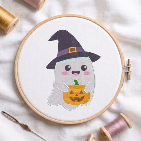 A circular embroidery hoop holding a white fabric with a cute embroidered ghost character wearing a black witch hat and holding a yellow-orange jack-o'-lantern. The hoop is styled on a white fabric background with spools of thread and a needle.