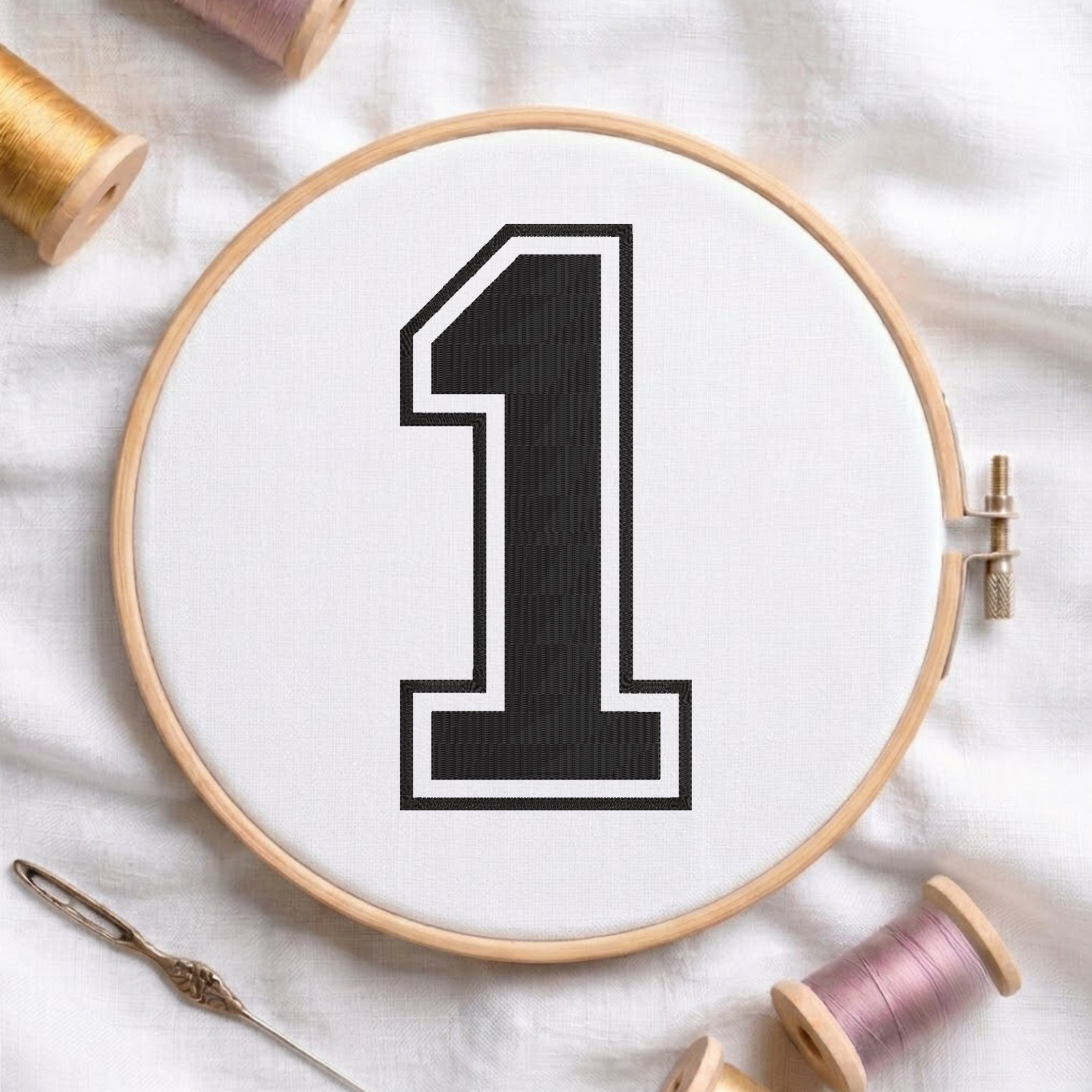 A wooden embroidery hoop holding white fabric with a large black number 1 outlined in white. A needle lies at the lower left, and spools of thread are partially visible around the hoop on a white fabric background.