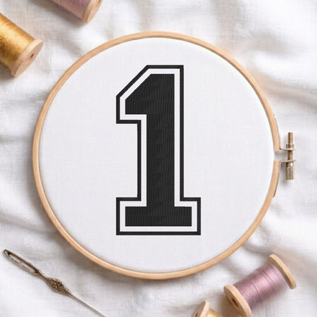 A wooden embroidery hoop holding white fabric with a large black number 1 outlined in white. A needle lies at the lower left, and spools of thread are partially visible around the hoop on a white fabric background.