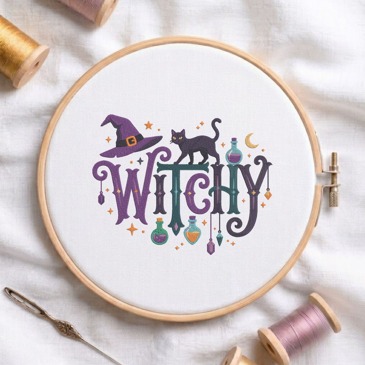 A circular embroidery hoop on white fabric, featuring a Halloween-themed cross-stitch design with the word “WITCH” in purple and dark lettering, a black cat, a witch hat, stars, potion bottles, and a crescent moon. Spools of thread, a needle, and a wooden stitch comb are arranged around the hoop.