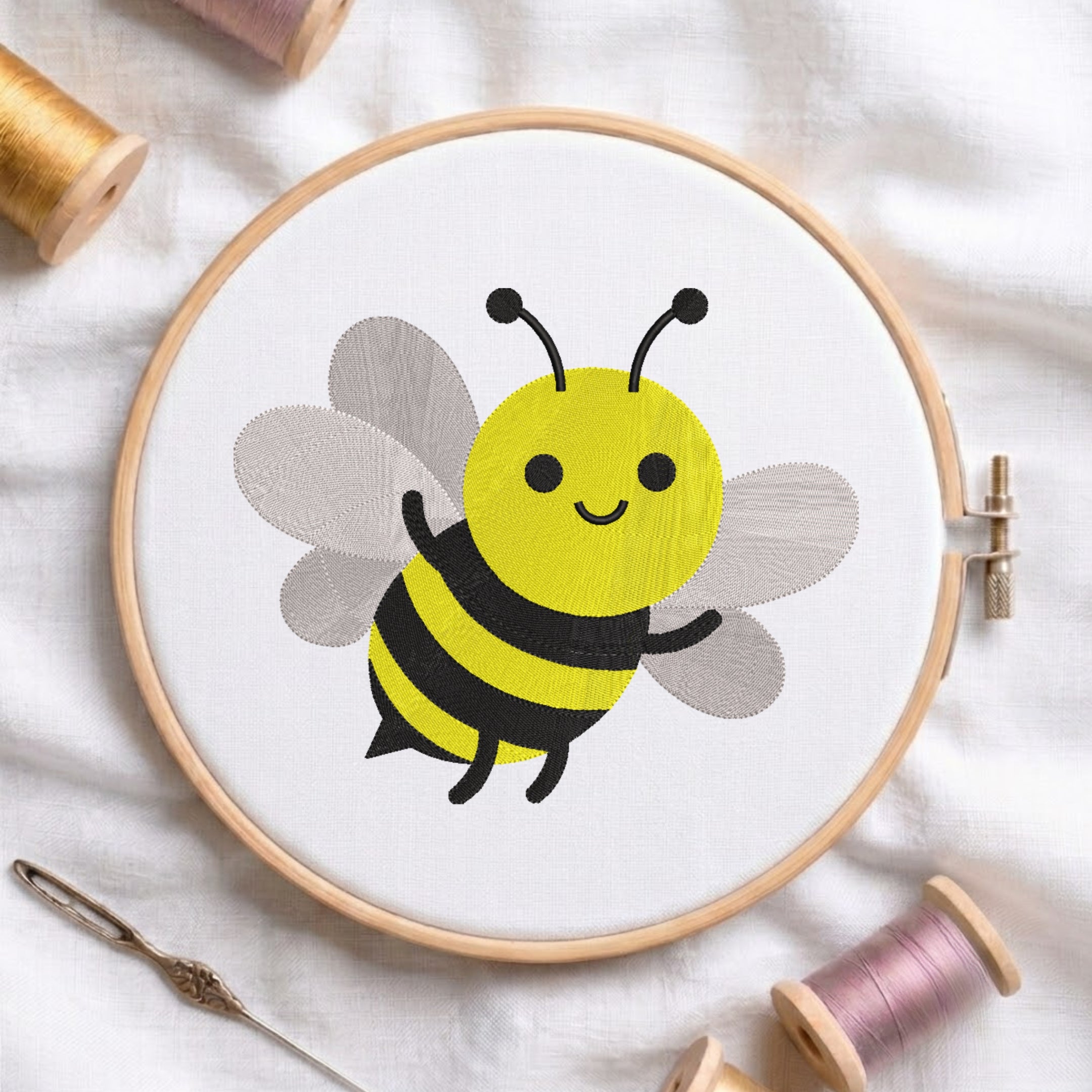A wooden embroidery hoop holding white fabric with a cartoon bumblebee design in yellow, black, and gray. The hoop is styled on a white fabric background with sewing supplies including thread spools, a needle, and a pin.