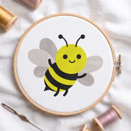 A wooden embroidery hoop holding white fabric with a cartoon bumblebee design in yellow, black, and gray. The hoop is styled on a white fabric background with sewing supplies including thread spools, a needle, and a pin.