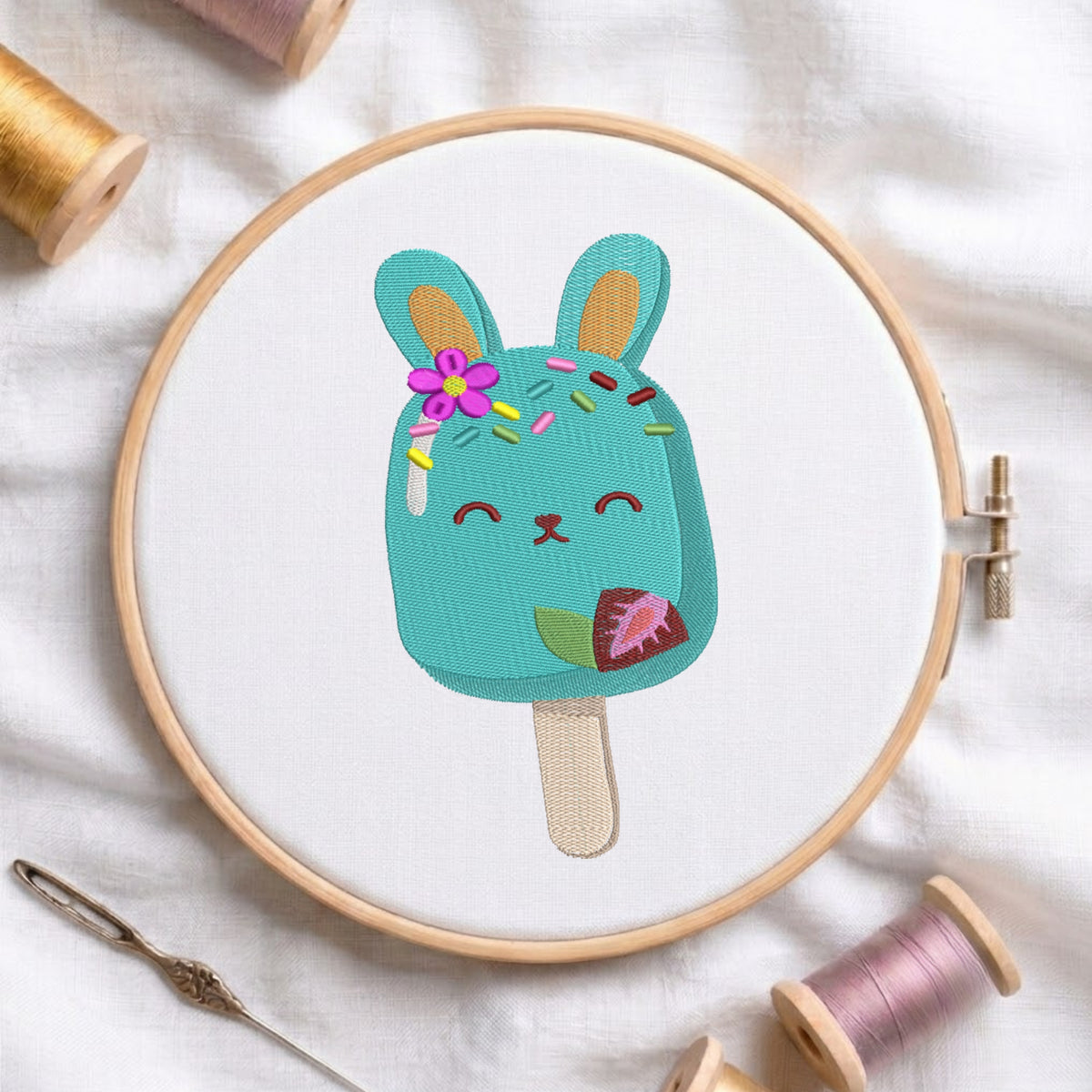 A round embroidery hoop holding a white fabric with a colorful embroidered bunny popsicle design. The design features a turquoise popsicle body with a pink flower, rainbow sprinkles, closed smiling eyes, and a small green leaf on the side.