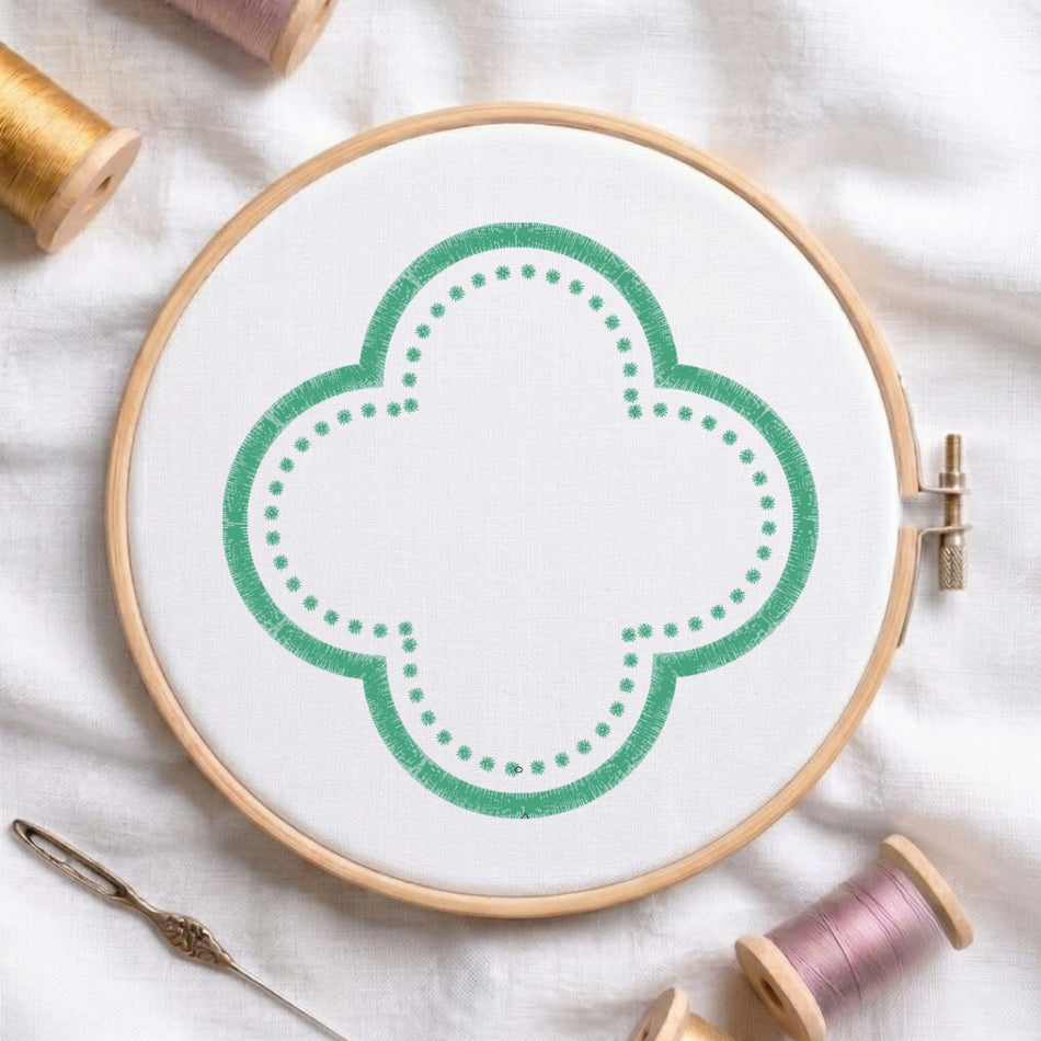 A wooden embroidery hoop holding white fabric with a green quatrefoil-style stitched design, surrounded by spools of thread and a needle on a white fabric background.