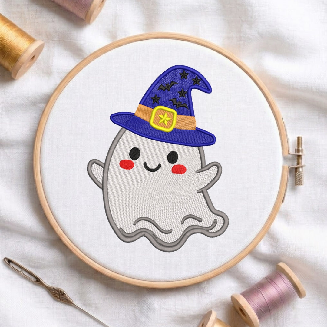 A circular embroidery hoop holding white fabric with a cute embroidered ghost wearing a purple wizard hat with black stars and a yellow band. The hoop is styled on a white fabric background with spools of thread and a needle.