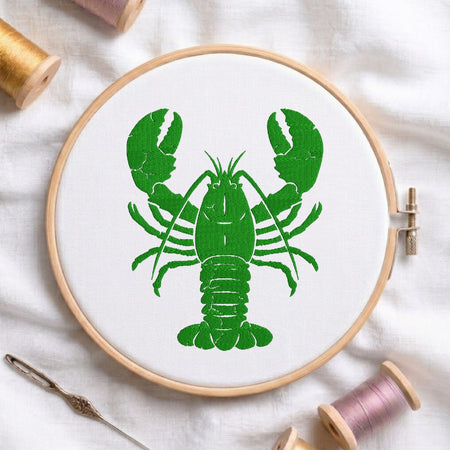A circular embroidery hoop holding white fabric with a bright green lobster design, styled on a white cloth with thread spools and a needle around it.