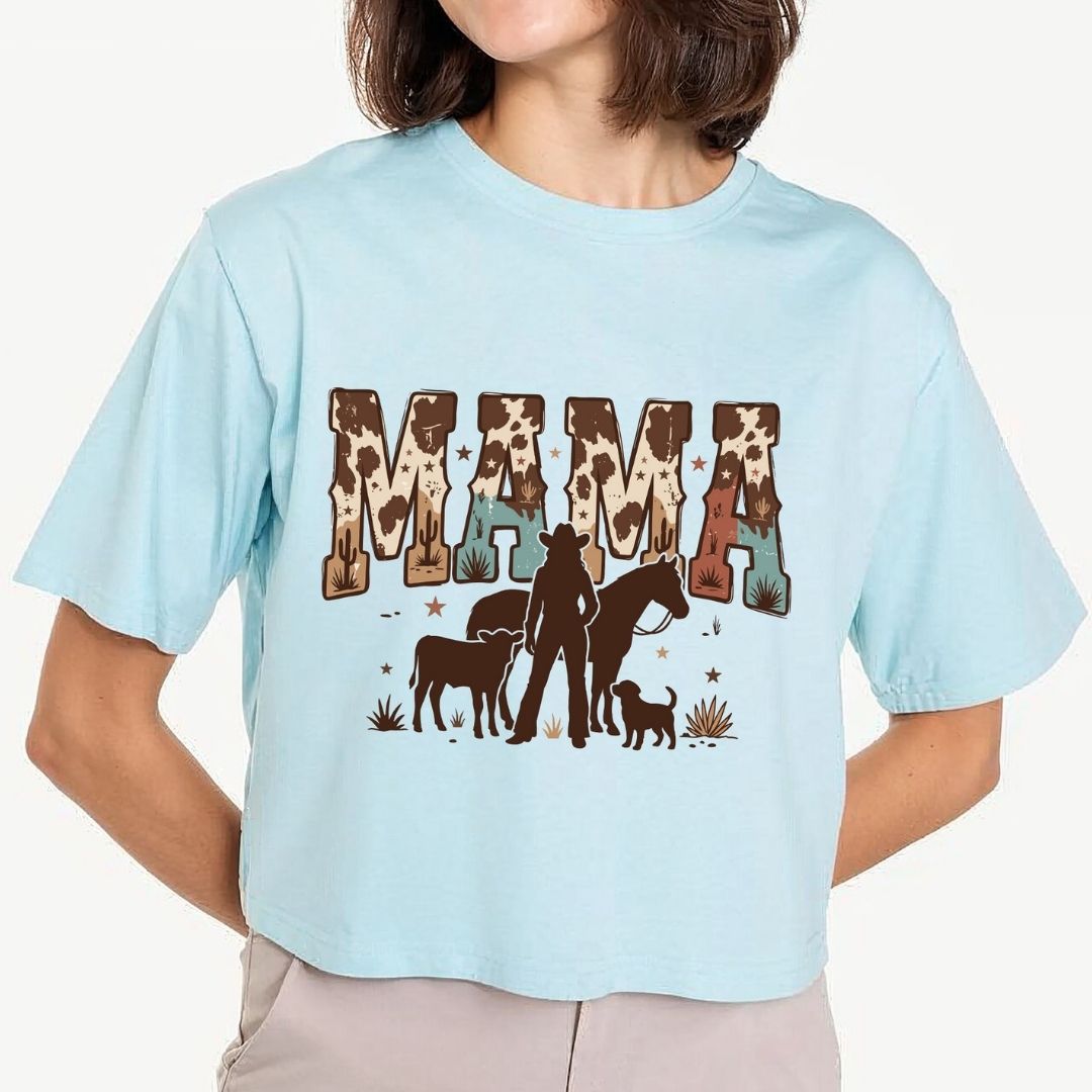 A woman wearing a light blue cropped t-shirt with a graphic that reads 'MAMA' in cow-print letters, with a silhouette of a woman, horse, and dog underneath.