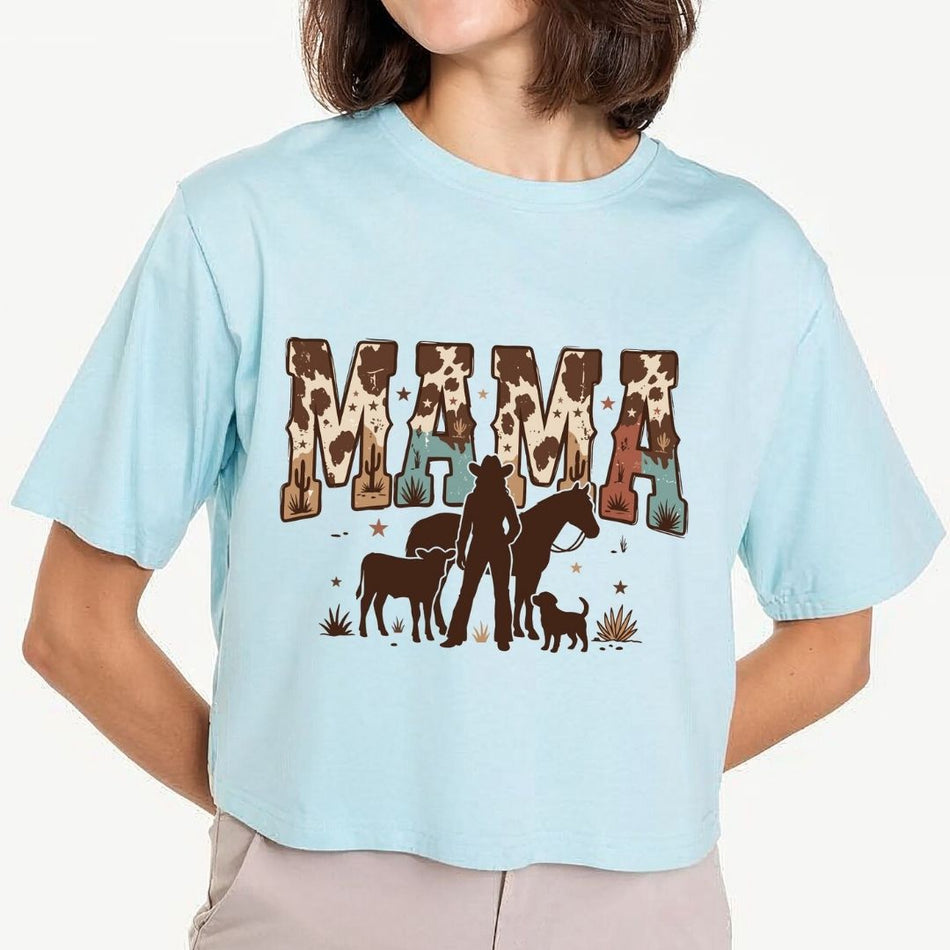 A woman wearing a light blue cropped t-shirt with a graphic that reads 'MAMA' in cow-print letters, with a silhouette of a woman, horse, and dog underneath.