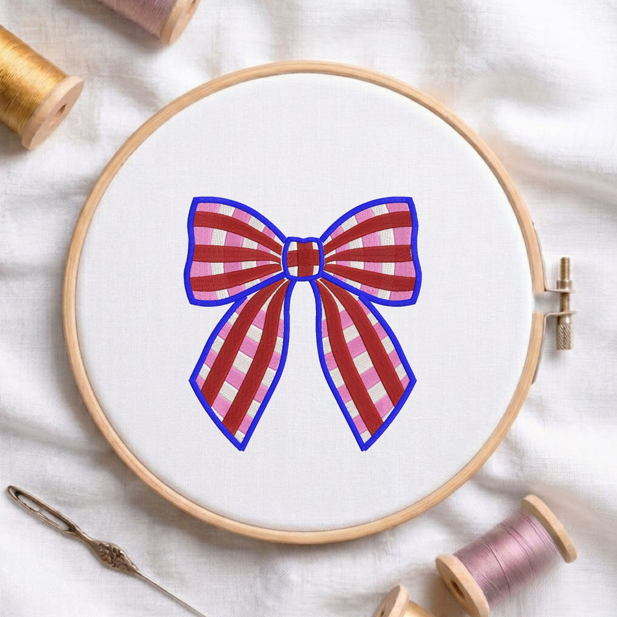 Red and pink plaid bow machine embroidery design displayed inside a round wooden hoop on white fabric alongside thread spools and a needle.