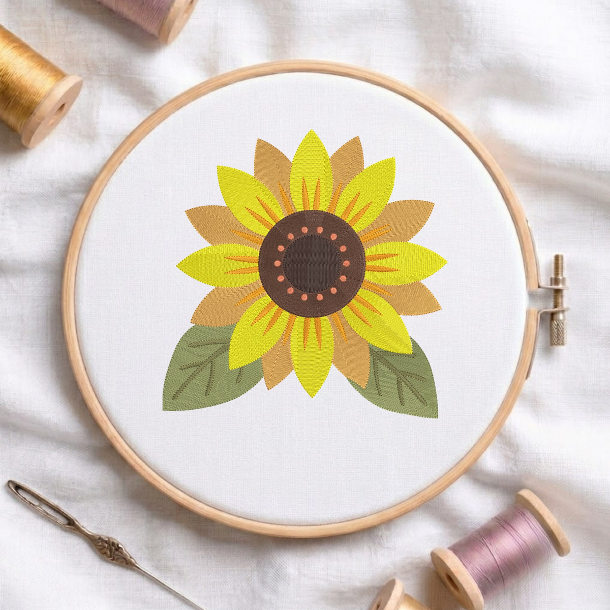 A wooden embroidery hoop holds white fabric with a stylized sunflower design in yellow, tan, green, and brown. The hoop is photographed on a white fabric surface with thread spools, a needle, and a pin around it.