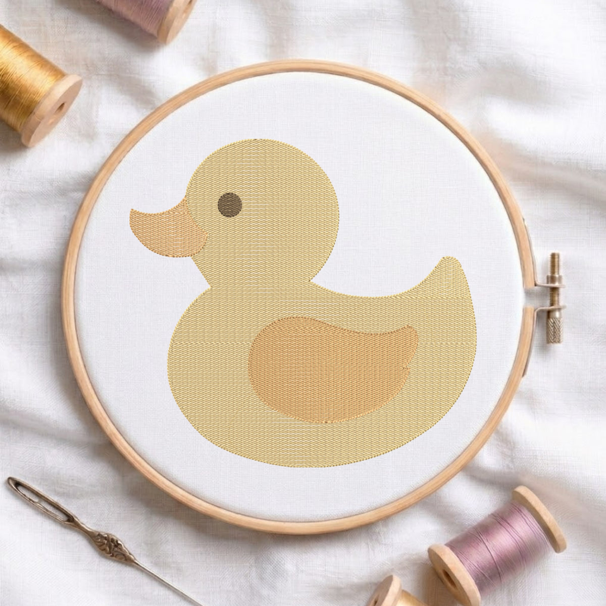 A wooden embroidery hoop holding white fabric with a stylized yellow duck design, surrounded by spools of thread and a needle on a soft white fabric background.