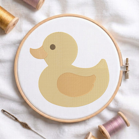 A wooden embroidery hoop holding white fabric with a stylized yellow duck design, surrounded by spools of thread and a needle on a soft white fabric background.