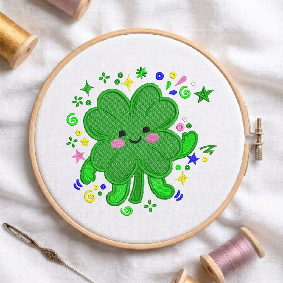 A circular embroidery hoop holding white fabric with a colorful embroidered green clover character surrounded by small stars, flowers, and swirls. Spools of thread and a needle are arranged around the hoop on a white fabric background.
