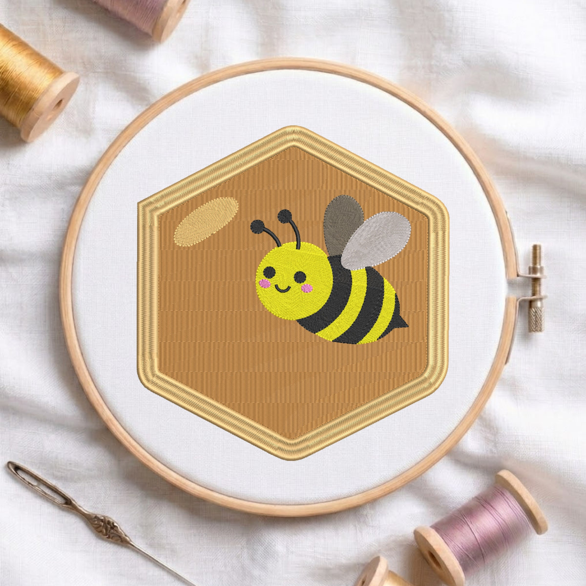 A wooden embroidery hoop holding a stitched design of a yellow and black bumblebee on a tan hexagonal background, surrounded by spools of thread and a needle on white fabric.