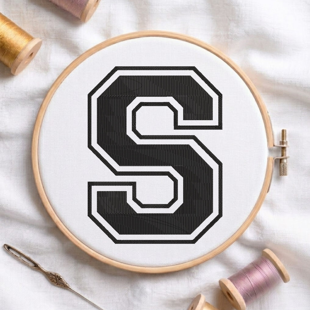 Close-up view of an embroidered varsity-style block letter 'S' in black with a white and black outline, centered in a wooden embroidery hoop surrounded by thread spools and a craft needle.