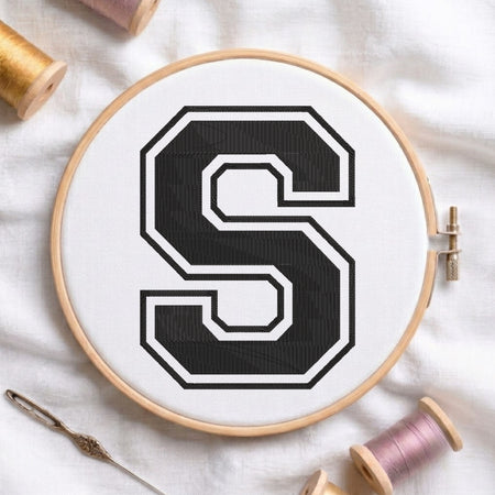 Close-up view of an embroidered varsity-style block letter 'S' in black with a white and black outline, centered in a wooden embroidery hoop surrounded by thread spools and a craft needle.