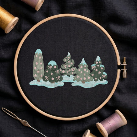 A circular embroidery hoop holds a black fabric piece with a stitched winter tree scene in mint, teal, and pink. The hoop is photographed on dark fabric with thread spools and a needle around it.