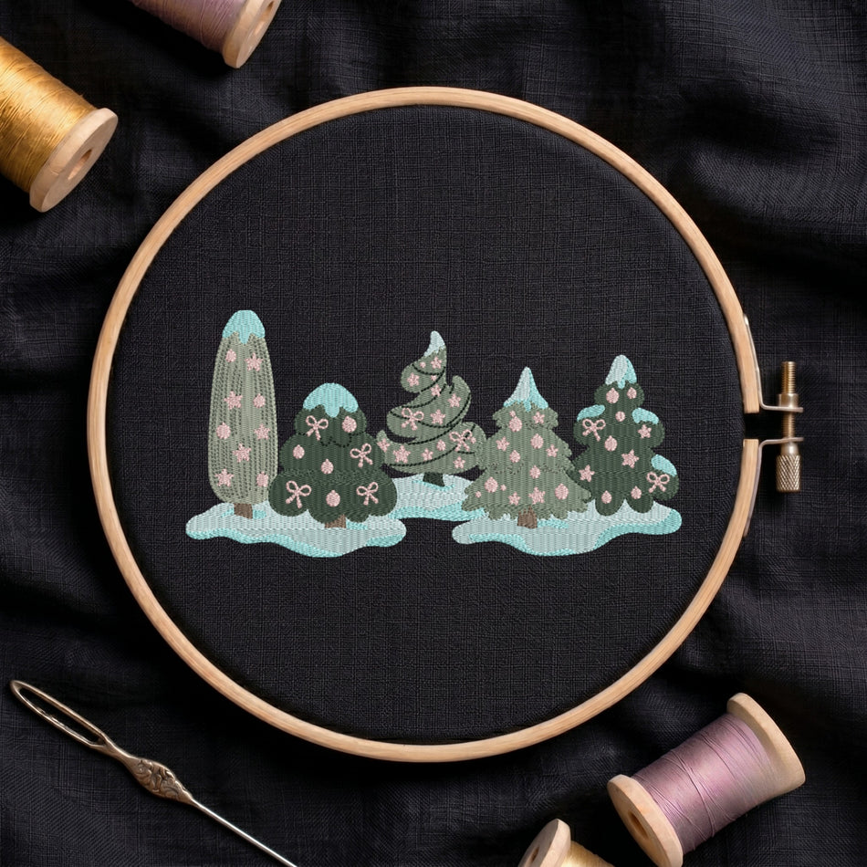 A circular embroidery hoop holds a black fabric piece with a stitched winter tree scene in mint, teal, and pink. The hoop is photographed on dark fabric with thread spools and a needle around it.