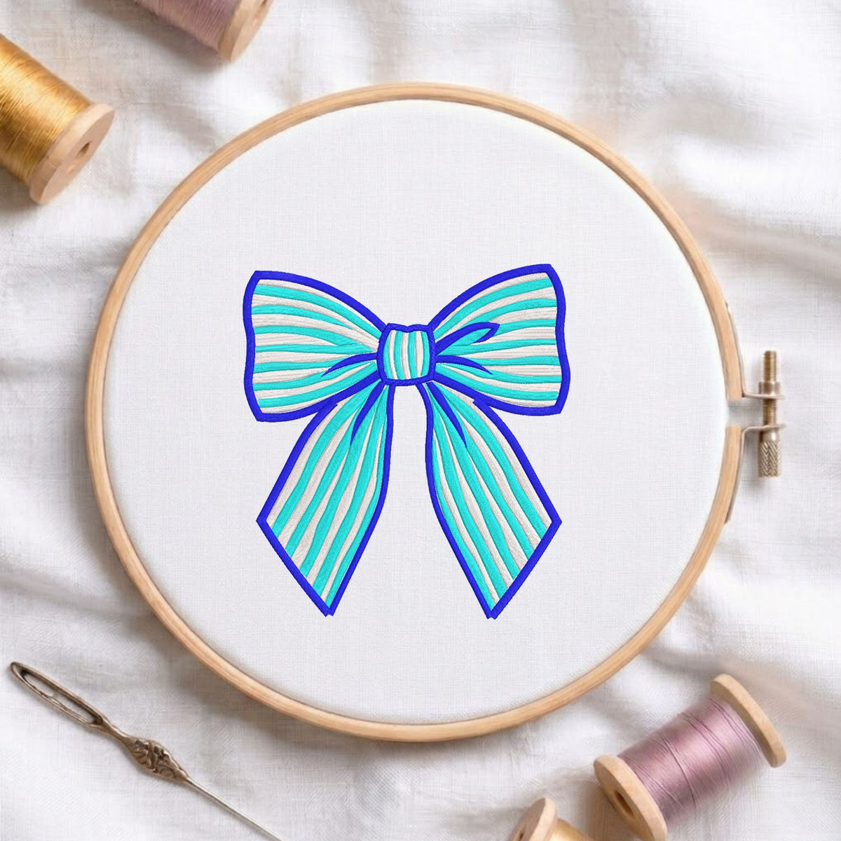 A wooden embroidery hoop holding white fabric with a stylized bow design in turquoise and blue, surrounded by spools of thread and a needle on a white fabric background.