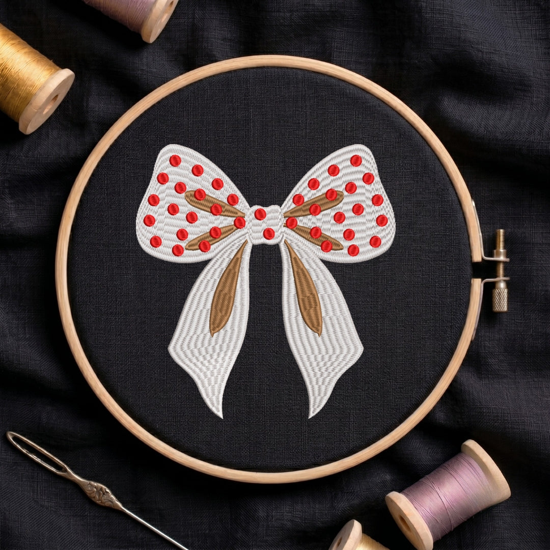 A circular embroidery hoop holding black fabric with a bow design stitched in white, red, and brown thread, surrounded by spools of thread and a needle on a dark fabric background.