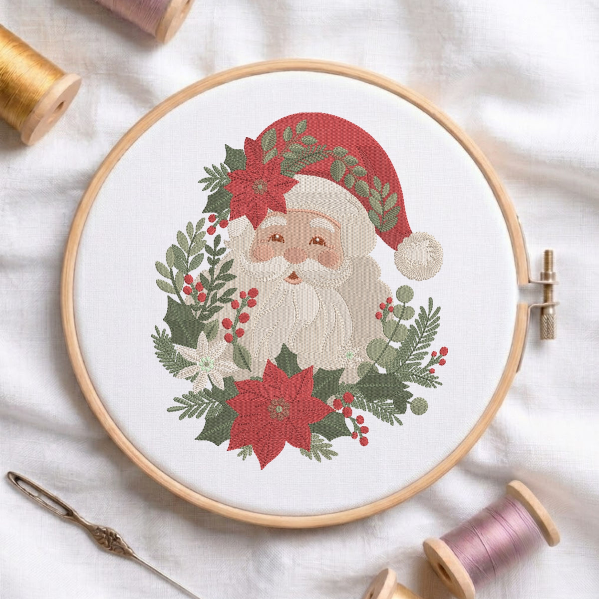 A circular embroidery hoop holds a festive Santa Claus design surrounded by poinsettias, holly leaves, and berries on white fabric. A needle lies at the lower left, and spools of thread are arranged around the hoop.