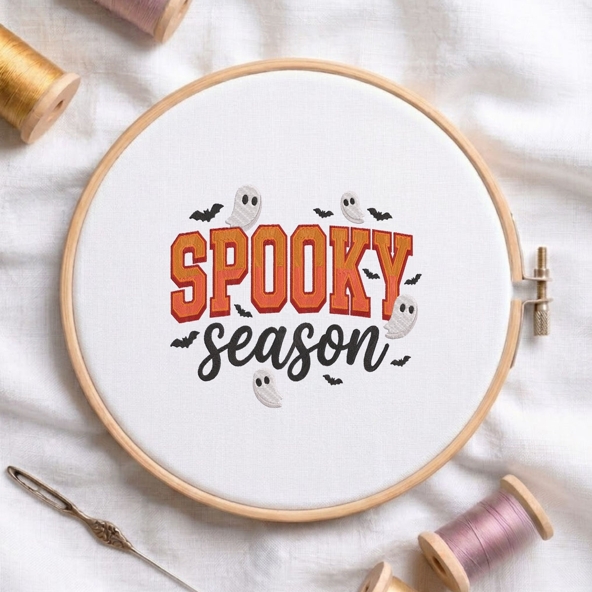 Cute spooky season lettering design featuring little white ghosts and black bats machine embroidery, displayed inside a round wooden hoop on white fabric with thread spools and a needle.
