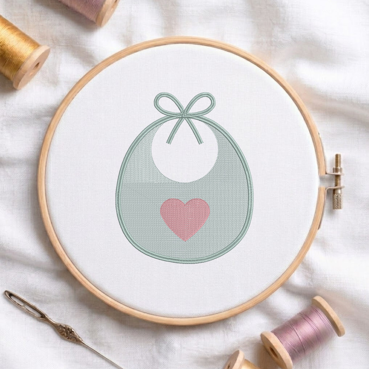 A circular embroidery hoop holding white fabric with a stitched design of a white bib or pouch outlined in pale green and accented with a pink heart. Spools of thread, a needle, and soft fabric are arranged around it.