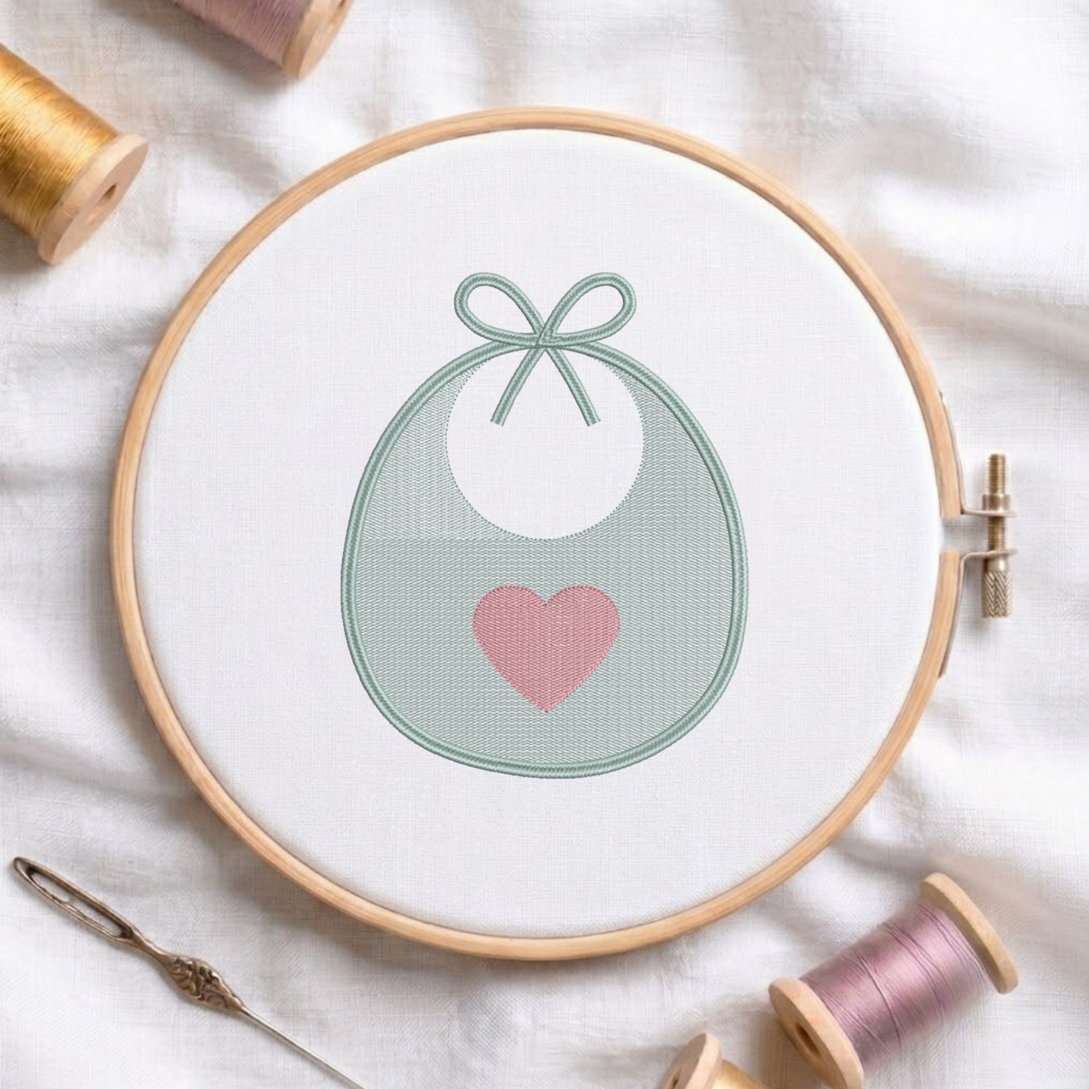 A circular embroidery hoop holding white fabric with a stitched design of a white bib or pouch outlined in pale green and accented with a pink heart. Spools of thread, a needle, and soft fabric are arranged around it.