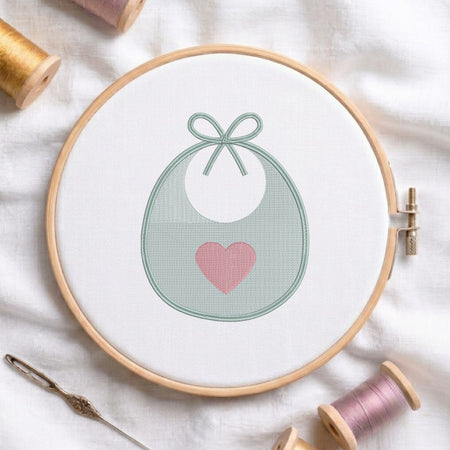 A circular embroidery hoop holding white fabric with a stitched design of a white bib or pouch outlined in pale green and accented with a pink heart. Spools of thread, a needle, and soft fabric are arranged around it.