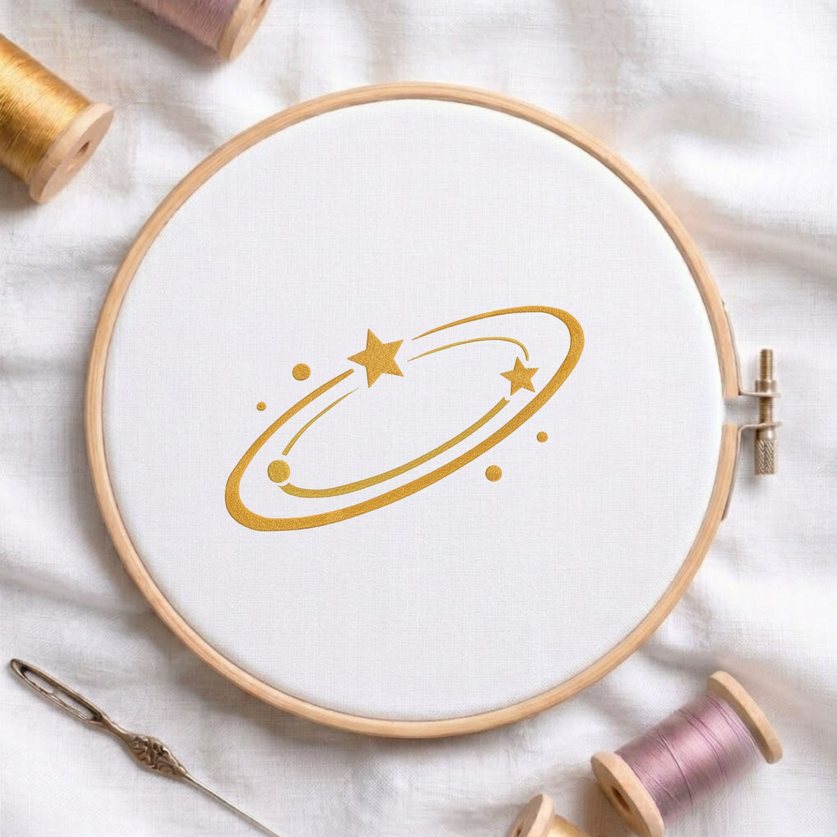 A circular embroidery hoop holding white fabric, with a gold celestial design of stars and rings stitched in the center. The hoop is styled on a white fabric background with spools of thread and a needle nearby.
