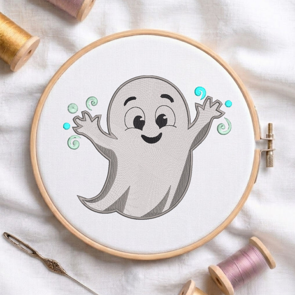 A circular embroidery hoop holding white fabric with a cartoon ghost design in gray, black, and light blue. The hoop is photographed on a white fabric surface with spools of thread and a needle around it.