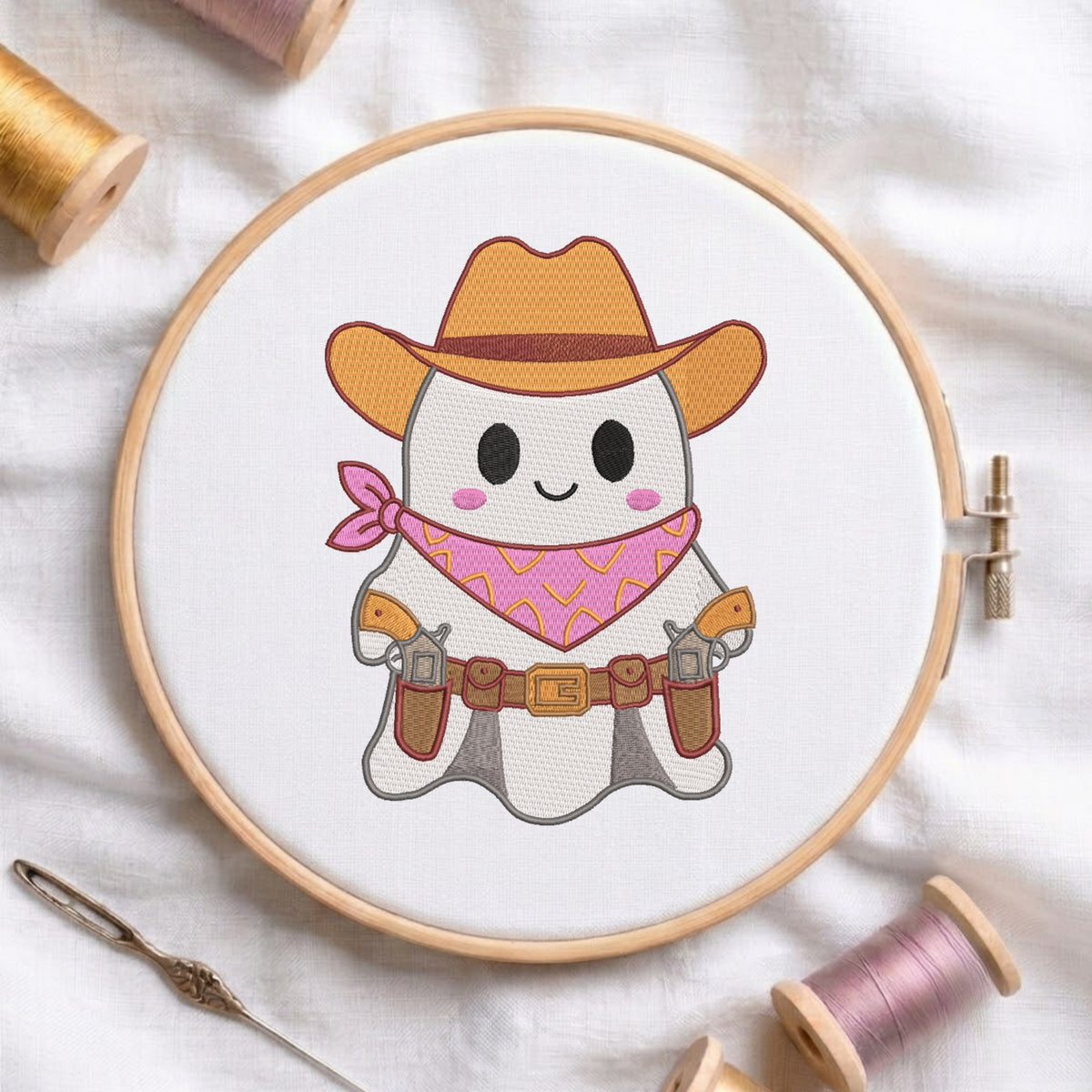 A white fabric stretched in a wooden embroidery hoop, featuring an embroidered design of a cute ghost wearing a cowboy hat, pink patterned bandana, and gun holsters.