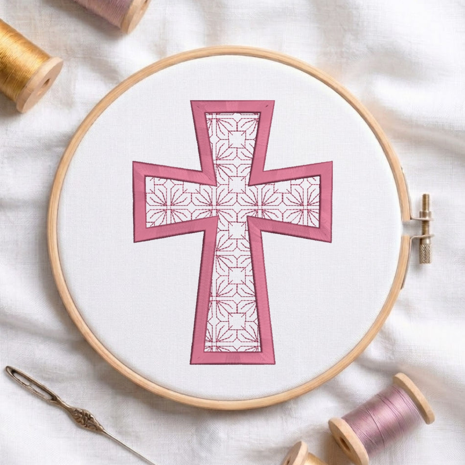 A circular embroidery hoop holding white fabric with a pink cross design, surrounded by spools of thread and a needle on a white fabric background.