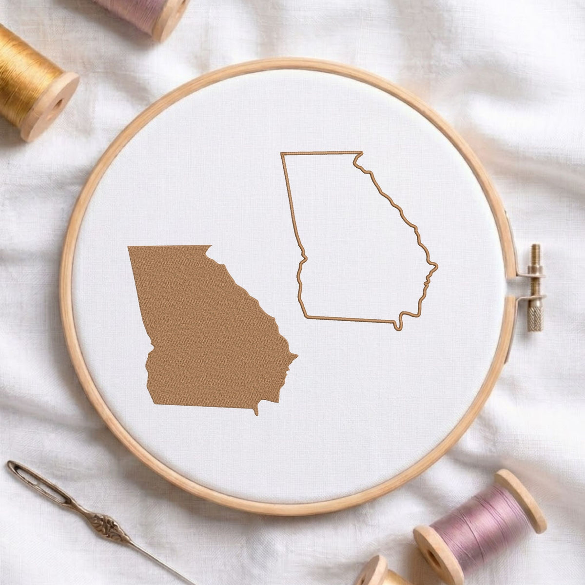 A circular embroidery hoop on white fabric, showing a Georgia state design in outline and filled with brown thread. Spools of thread and a needle are arranged around it on a soft white fabric background.